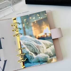 May include: A light pink leather binder with a gold six-ring mechanism. The interior features a divider with a printed image of a white knitted blanket and blurred golden lights. The text "CHAPTER 1 - HIGH PRIORITY" and "HONED" are visible.