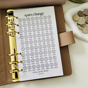 May include: A beige leather binder with gold rings holding a white paper savings tracker titled spare change. The tracker features rows of circular icons with dollar signs and incremental values, ending with a total saved amount of $131.40.