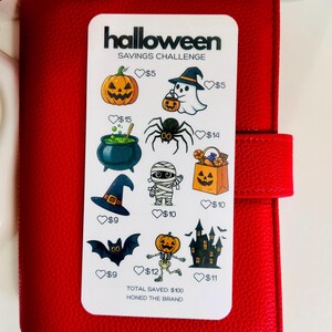 May include: A white Halloween savings challenge card with illustrations of pumpkins, ghosts, and other spooky images. The card is on a red leather-like surface. The text "Halloween Savings Challenge" is at the top.