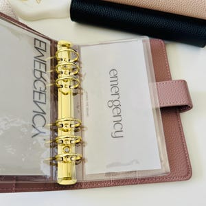 May include: A dusty rose textured leather binder featuring a gold-toned six-ring metal mechanism. The interior contains clear plastic dividers with the word emergency printed in black text, along with the smaller text HONED THE BRAND.