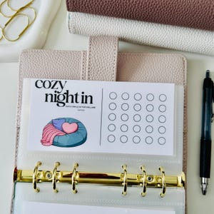 Può includere: Un raccoglitore beige a sei anelli contenente una scheda bianca per il monitoraggio dei risparmi con il testo "cozy night in" e "each circle is five dollars". La scheda presenta un pouf blu, una coperta rosa e un cuore, insieme a venticinque cerchi vuoti.