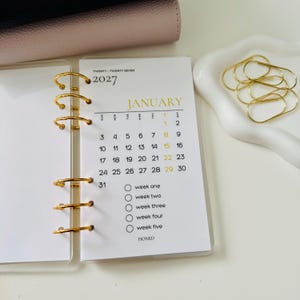 Peut inclure: Une page d'insertion de planificateur blanche pour janvier 2027 avec une reliure à anneaux dorés. La page présente une grille de calendrier mensuel, le texte TWENTY-TWENTY SEVEN, JANUARY, et une liste avec cinq cercles nommés week one à week five et le mot HONED.