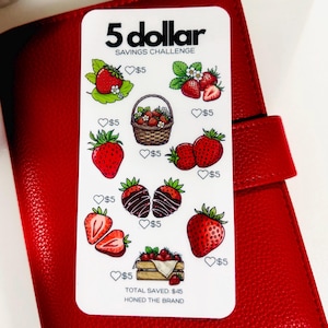 May include: A white savings challenge card with the text "5 dollar SAVINGS CHALLENGE" and illustrations of strawberries. The card is on a red leather surface. Each strawberry illustration has a £5 symbol.