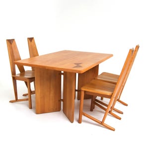May include: A light brown wooden dining table with four matching chairs. The table has a rectangular top with a decorative inlay and a sturdy base. The chairs have tall backs and a simple design.