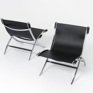 Pode incluir: Duas cadeiras de couro preto com estrutura cromada. As cadeiras têm um design minimalista, com encosto e assento curvos. As pernas são anguladas, proporcionando estabilidade e uma estética moderna, em fundo branco.