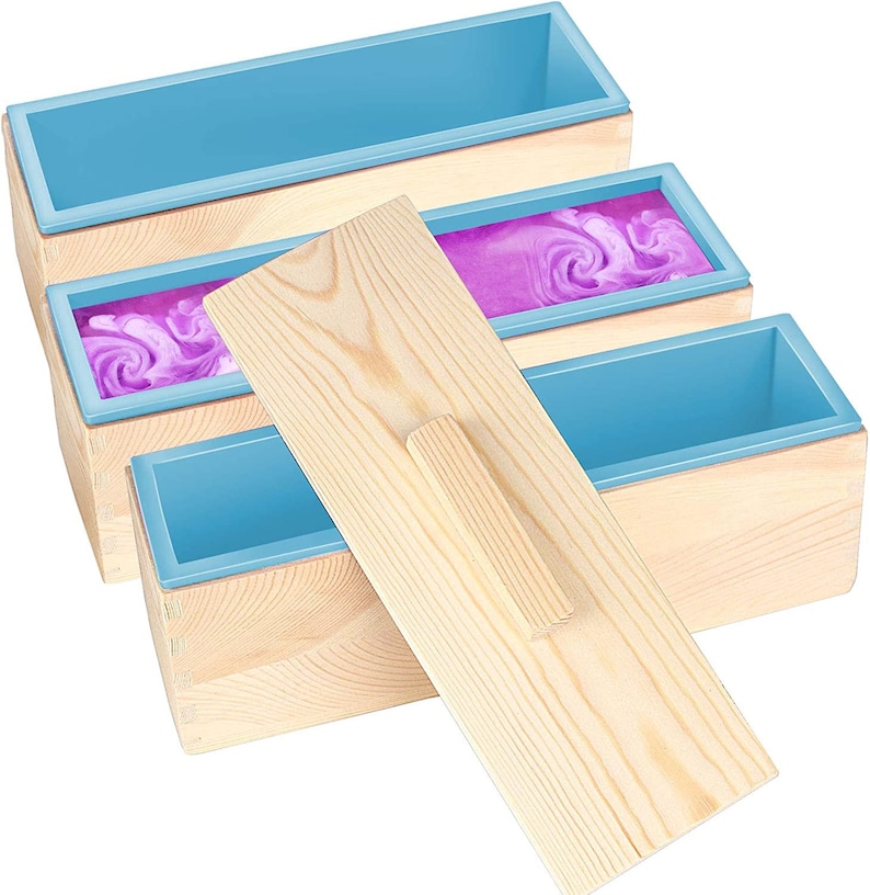 Pode incluir: Um conjunto de tr&ecirc;s moldes de sab&atilde;o de madeira com inser&ccedil;&otilde;es de silicone azul. Um molde cont&eacute;m um redemoinho de sab&atilde;o roxo. Uma ferramenta de alisamento de madeira repousa sobre os moldes. A madeira tem uma cor clara natural.