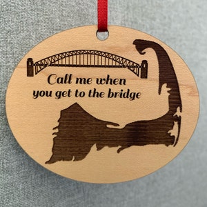 May include: A wooden ornament with a silhouette of the Cape Cod coastline and the Sydney Harbour Bridge. The ornament has the text "Call me when you get to the bridge" engraved on it.