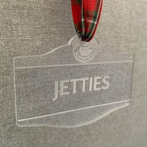 May include: A clear acrylic ornament with a red and green plaid ribbon. The ornament has a raised edge and the word "JETTIES" is engraved on the front.