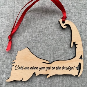 May include: A wooden ornament in the shape of a geographical outline, with a red ribbon for hanging. The ornament has the text "Call me when you get to the bridge!" etched into the wood.