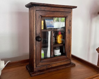 May include: A dark brown wooden cabinet with a glass door and shelves. The cabinet has a small knob on the door and is filled with various items, including a bottle of liquid, a box of bandages, and a white container.
