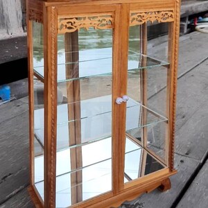 Puede incluir: Una vitrina de madera con puertas y estantes de vidrio. La vitrina tiene tallas intrincadas en la parte delantera y los lados. La vitrina est&aacute; hecha de madera marr&oacute;n claro y tiene una tapa de vidrio transparente.