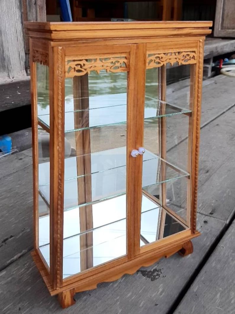 Puede incluir: Una vitrina de madera con puertas y estantes de vidrio. La vitrina tiene tallas intrincadas en la parte delantera y los lados. La vitrina est&aacute; hecha de madera marr&oacute;n claro y tiene una tapa de vidrio transparente.