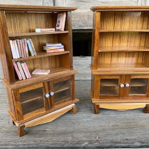 Op de afbeelding: Twee miniatuur houten boekenkasten met glazen deuren en planken. De boekenkast aan de linkerkant heeft boeken op de planken. Beide boekenkasten hebben een houtnerf afwerking.