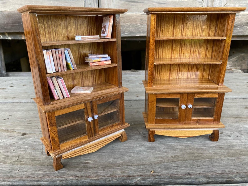 Op de afbeelding: Twee miniatuur houten boekenkasten met glazen deuren en planken. De boekenkast aan de linkerkant heeft boeken op de planken. Beide boekenkasten hebben een houtnerf afwerking.