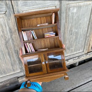 Op de afbeelding: Een miniatuur houten boekenkast met glazen deuren en planken gevuld met miniatuur boeken. De boekenkast is bruin met een houtnerf.