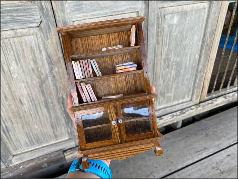 Op de afbeelding: Een miniatuur houten boekenkast met glazen deuren en planken gevuld met miniatuur boeken. De boekenkast is bruin met een houtnerf.