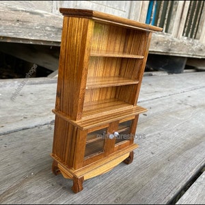 Op de afbeelding: Een miniatuur houten kast met twee glazen deuren en drie planken. De kast is gemaakt van lichtbruin hout met een gestreept patroon. De kast heeft zilveren knoppen op de deuren.