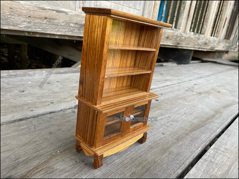 Op de afbeelding: Een miniatuur houten kast met twee glazen deuren en drie planken. De kast is gemaakt van lichtbruin hout met een gestreept patroon. De kast heeft zilveren knoppen op de deuren.