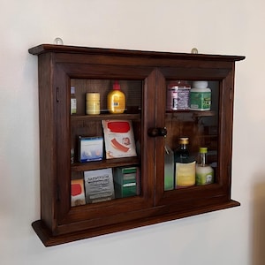 May include: A wall-mounted dark brown wooden cabinet with two glass-paneled doors and round wooden knobs. The interior features two shelves holding various medicine bottles, boxes, and containers, including a yellow bottle and white boxes.