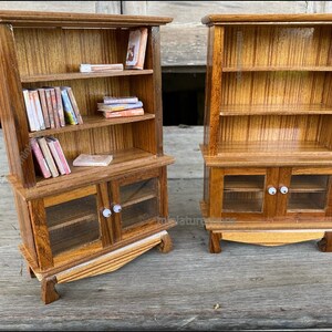 Op de afbeelding: Twee miniatuurhouten boekenkasten met glazen deuren. De boekenkast links heeft drie planken en is gevuld met miniatuurboeken. De boekenkast rechts heeft twee planken en is leeg.