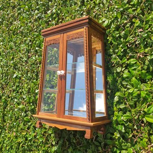 Puede incluir: Una vitrina de madera con puertas y estantes de vidrio. La vitrina tiene tallas intrincadas y un acabado marr&oacute;n oscuro. Est&aacute; montada en una pared.