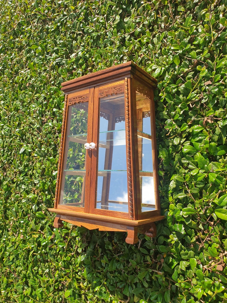 Puede incluir: Una vitrina de madera con puertas y estantes de vidrio. La vitrina tiene tallas intrincadas y un acabado marr&oacute;n oscuro. Est&aacute; montada en una pared.