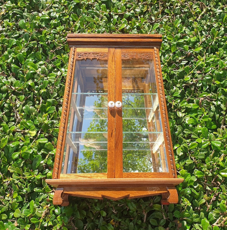 Puede incluir: Una vitrina de madera con puertas y estantes de vidrio. La vitrina tiene tallas intrincadas y es de un color marr&oacute;n intenso.