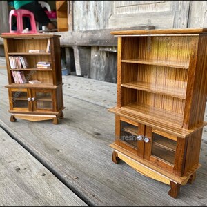 Op de afbeelding: Twee miniatuur houten boekenkasten met glazen deuren en planken. De boekenkasten zijn gemaakt van lichtbruin hout met een gestreept patroon. De bovenste boekenkast heeft boeken op de planken. De onderste boekenkast heeft glazen deuren met een kleine knop op elke deur.