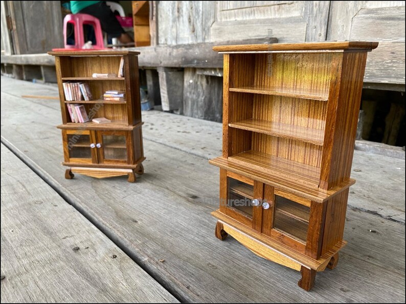 Op de afbeelding: Twee miniatuur houten boekenkasten met glazen deuren en planken. De boekenkasten zijn gemaakt van lichtbruin hout met een gestreept patroon. De bovenste boekenkast heeft boeken op de planken. De onderste boekenkast heeft glazen deuren met een kleine knop op elke deur.