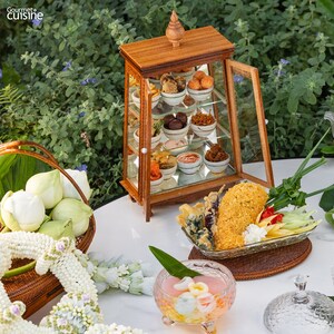 Puede incluir: Una vitrina de madera con varios platos peque&ntilde;os llenos de diferentes aperitivos. La vitrina est&aacute; sobre una mesa blanca con un cuenco de postre rosa, un plato de comida frita y una cesta de flores blancas.