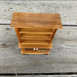 Op de afbeelding: Een houten hutch met een bovenste plank en twee onderste planken. De hutch heeft een glazen deur aan de onderkant en twee kleine witte knoppen.