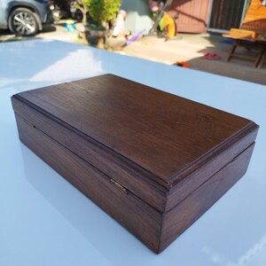 May include: A dark brown wooden box with a hinged lid and a brass clasp. The box is closed and sitting on a white surface.