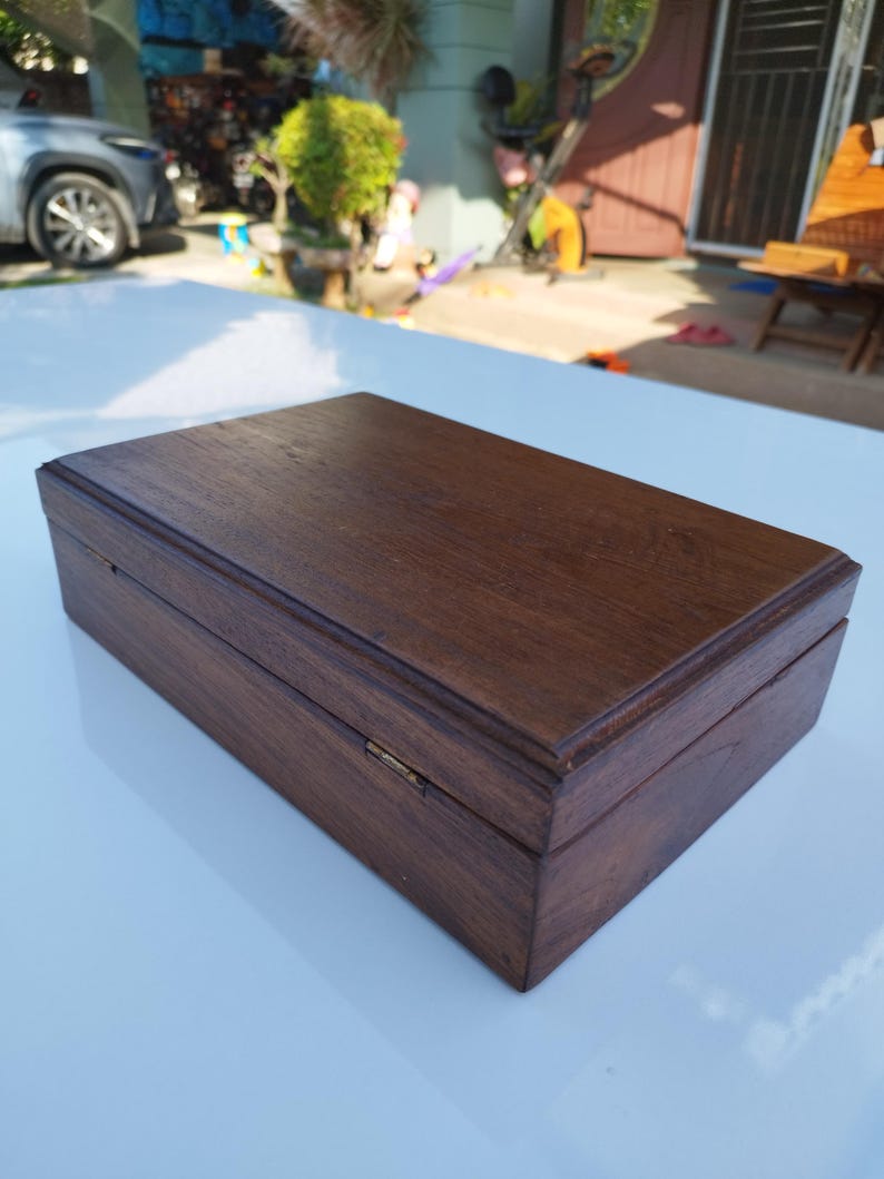 May include: A dark brown wooden box with a hinged lid and a brass clasp. The box is closed and sitting on a white surface.