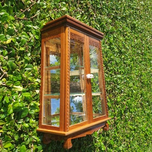 Puede incluir: Una vitrina de pared de madera con puertas y estantes de vidrio. La vitrina tiene tallas intrincadas y un acabado marr&oacute;n claro.