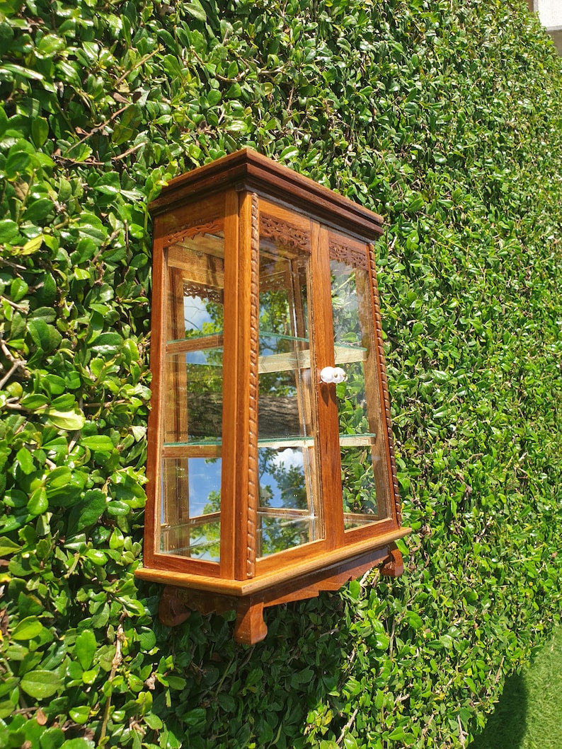 Puede incluir: Una vitrina de pared de madera con puertas y estantes de vidrio. La vitrina tiene tallas intrincadas y un acabado marr&oacute;n claro.