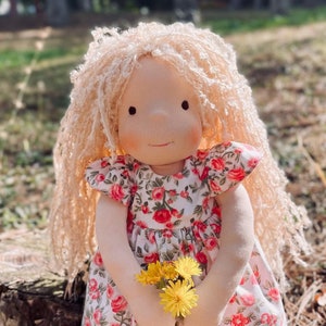 May include: A doll with long blonde hair wearing a floral dress and holding a bunch of yellow flowers. The doll is standing on a tree stump in a grassy area.