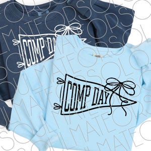 May include: A light blue crewneck sweatshirt and a dark navy blue crewneck sweatshirt. Each garment features a graphic of a pennant flag with a bow and the text COMP DAY printed in a bold, block-style font across the center of the chest area.