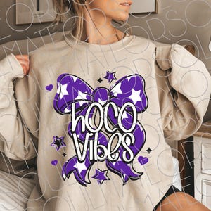May include: Beige sweatshirt with a purple bow graphic featuring white stars and the words "HOCO Vibes" in white. The design includes purple stars and hearts. A casual, comfortable top.