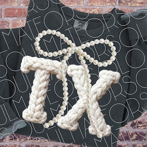 May include: Dark gray sweatshirt with a white knitted "TX" design, adorned with a pearl necklace in a bow shape. The letters are thick and textured, creating a unique visual contrast against the smooth fabric.