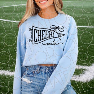 May include: A light blue sweatshirt with the words "CHEER coach" in black script and a bow graphic. The sweatshirt is a long-sleeved crew neck style. The text is inside a pennant shape.