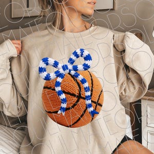May include: Beige sweatshirt featuring a basketball with a blue and white bow. The basketball is orange with black lines. The bow is made of a blue and white striped material. The sweatshirt is a neutral color.