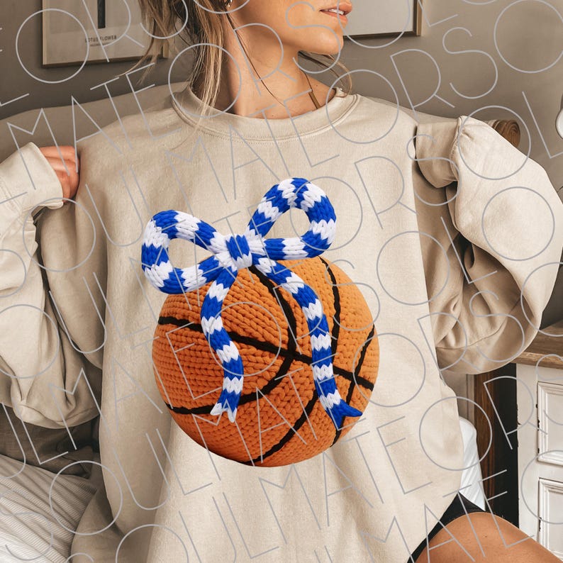 May include: Beige sweatshirt featuring a basketball with a blue and white bow. The basketball is orange with black lines. The bow is made of a blue and white striped material. The sweatshirt is a neutral color.