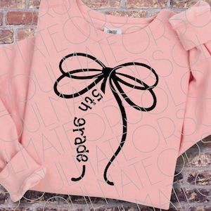 May include: A light peach-coloured long-sleeve sweatshirt featuring a large black graphic of a tied bow. One of the trailing ribbons of the bow contains the black text 5th grade written in a cursive font. The garment has a crew neckline.