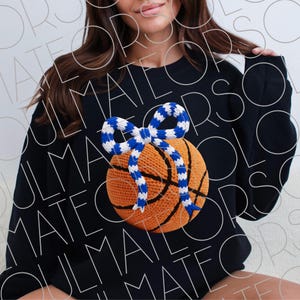 May include: Black sweatshirt featuring a basketball design with a blue and white bow. The basketball is orange with black lines, and the bow is a contrasting blue and white pattern. The sweatshirt is a casual style.