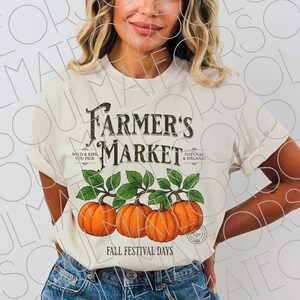 May include: Cream-colored t-shirt featuring the text "Farmer's Market" in a vintage style, with three orange pumpkins and green leaves. The shirt also includes the phrases "Wild & Ripe You Pick" and "Natural & Organic", and "Fall Festival Days".