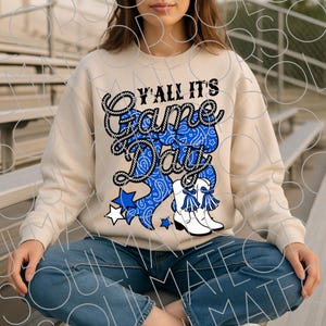 May include: A cream-colored sweatshirt with the text "Y'all It's Game Day" in black and blue. The design includes a blue bandana-style bow, white cowboy boots, and blue stars. The sweatshirt is a casual, comfortable style.