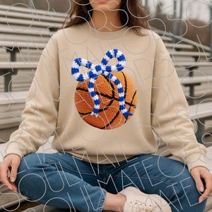 May include: A beige sweatshirt featuring a basketball design with a blue and white bow. The basketball is orange with black lines, and the bow is knitted. The sweatshirt is worn with blue jeans.