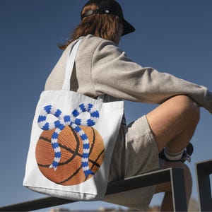 May include: White tote bag featuring a basketball design with a blue and white striped bow. The basketball is brown with black lines. The bag has white straps and is carried over the shoulder.