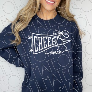 May include: Navy blue sweatshirt with a white pennant design that reads "CHEER coach". The pennant is decorated with a bow and small decorative elements. The sweatshirt is a crew neck style.