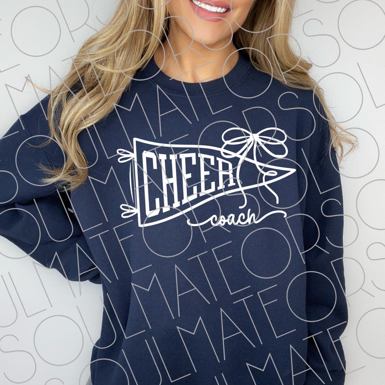 May include: Navy blue sweatshirt with a white pennant design that reads "CHEER coach". The pennant is decorated with a bow and small decorative elements. The sweatshirt is a crew neck style.